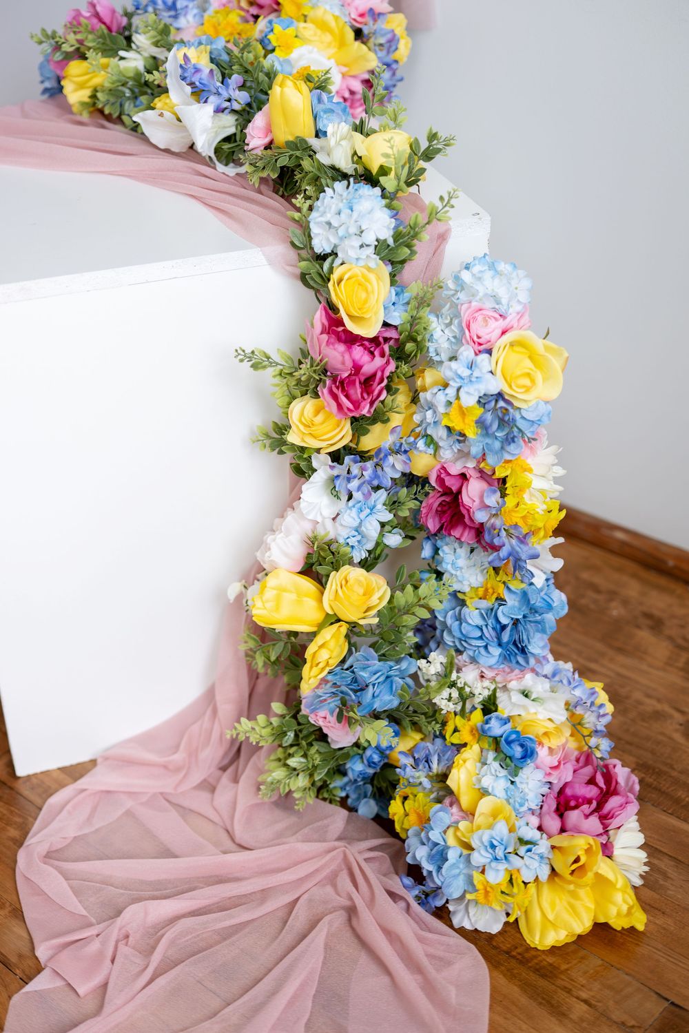 Vibrant floral garland with yellow roses, blue hydrangeas, and pink flowers cascading down white surface.