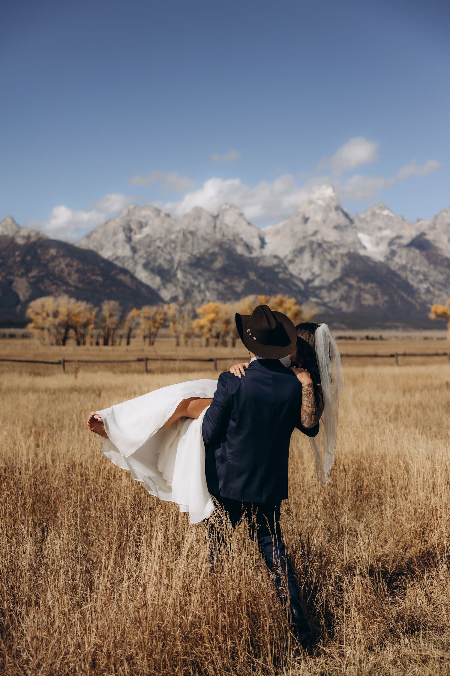 A bride in a flowing white dress poses playfully with her veil by a scenic mountain lake surrounded by rocky shores.