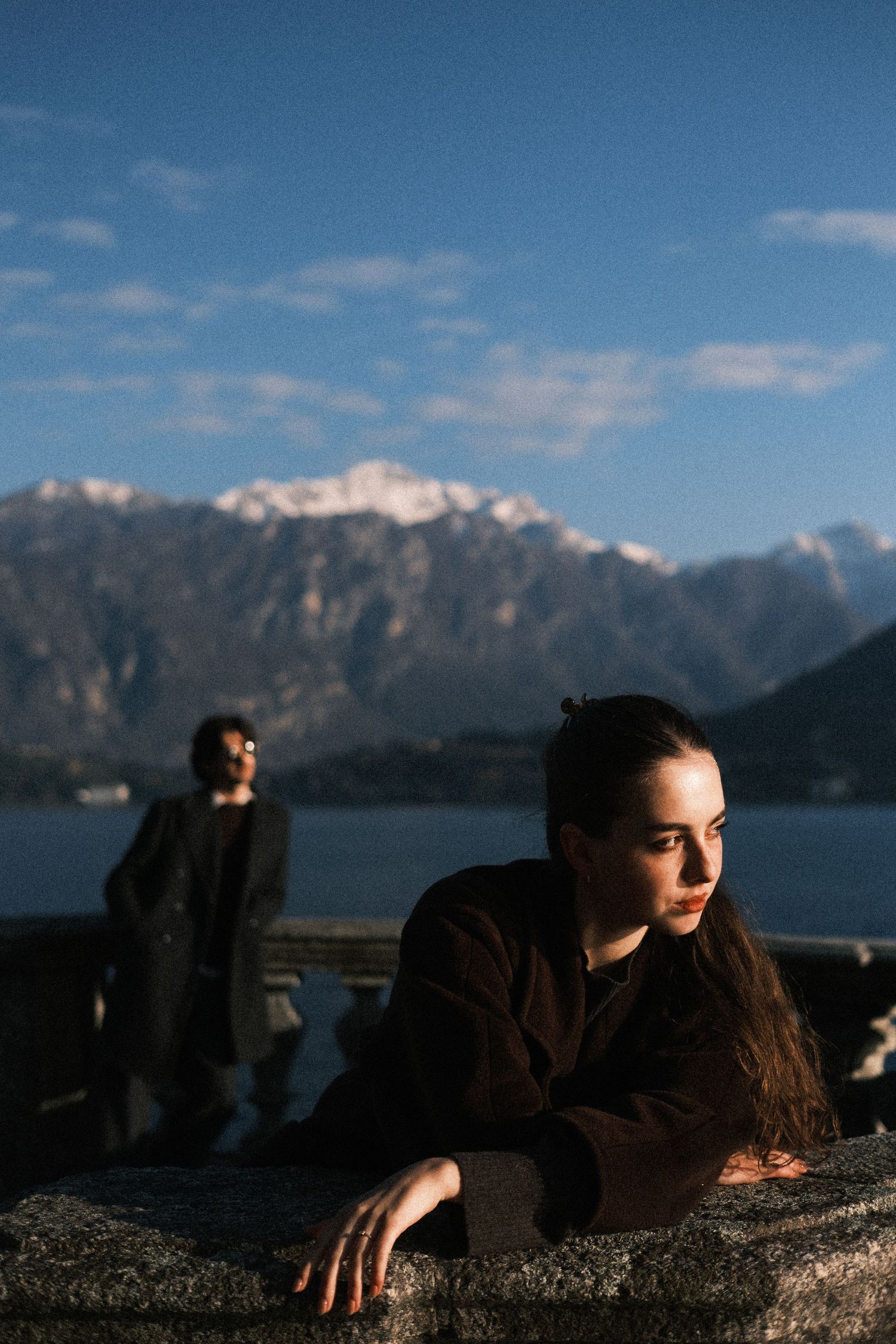 Silhouetted figures rest along a rocky ledge overlooking a scenic mountain lake on a sunny day.