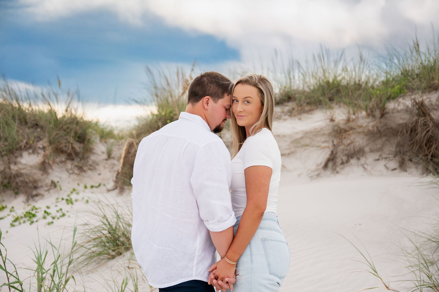 Romantic couple sharing an intimate moment on sandy beach dunes against blue sky.