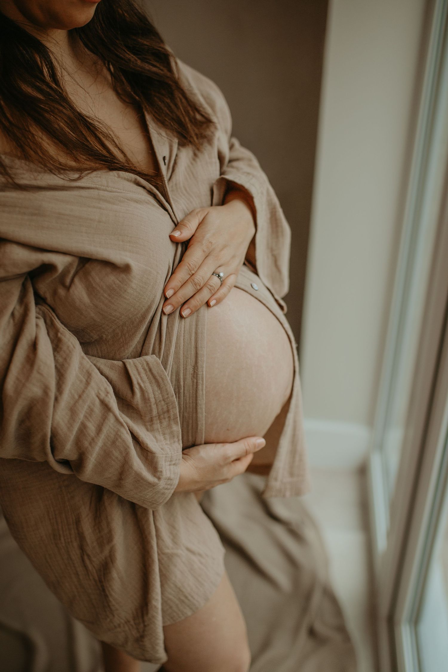 A dreamy maternity photography session featuring soft beige tones and natural light streaming through sheer curtains.