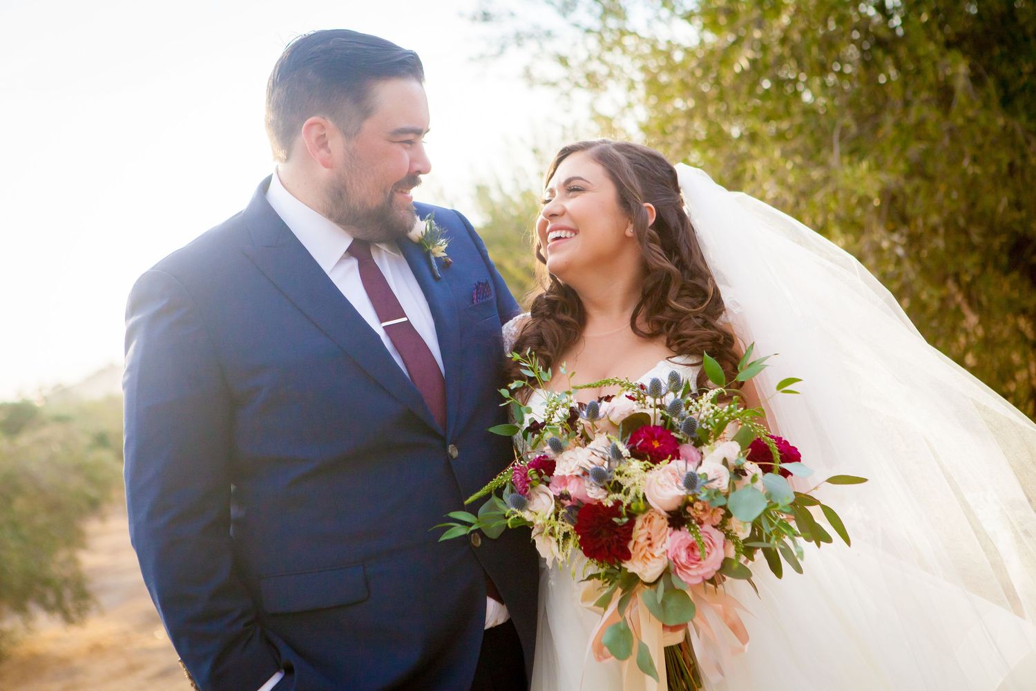 A couple shares a romantic moment outdoors during their wedding with burgundy floral bouquet and autumn scenery.