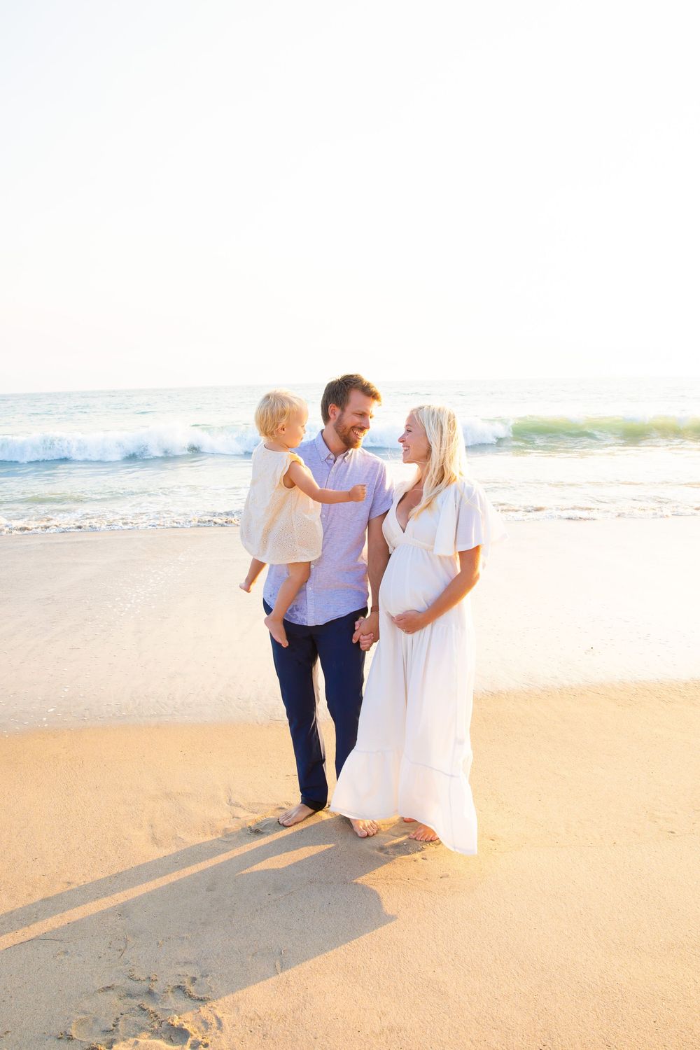 A couple walks hand in hand along a sandy beach at sunset, casting long shadows on the golden sand.