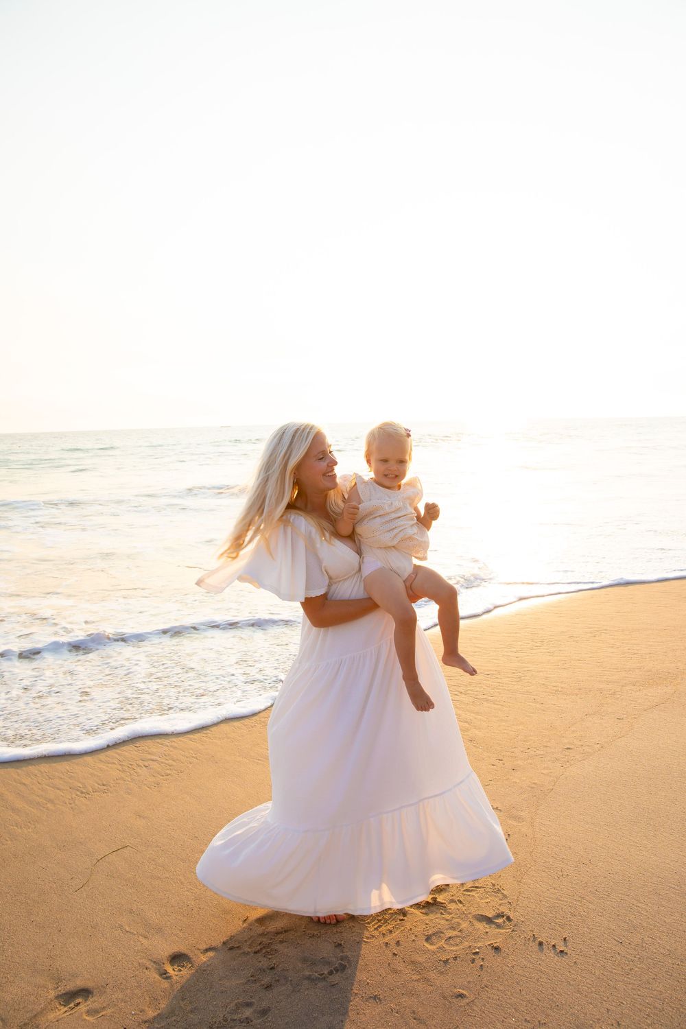 A mother in a white dress holds a baby while walking along a sandy beach at sunset.