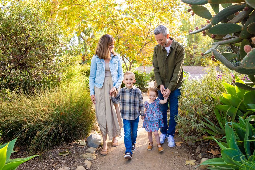 Family walking together through a garden path surrounded by autumn foliage and greenery in warm sunlight.