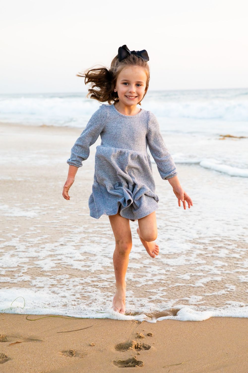 Child wearing gray romper jumps and plays in ocean waves at sandy beach during sunny day.