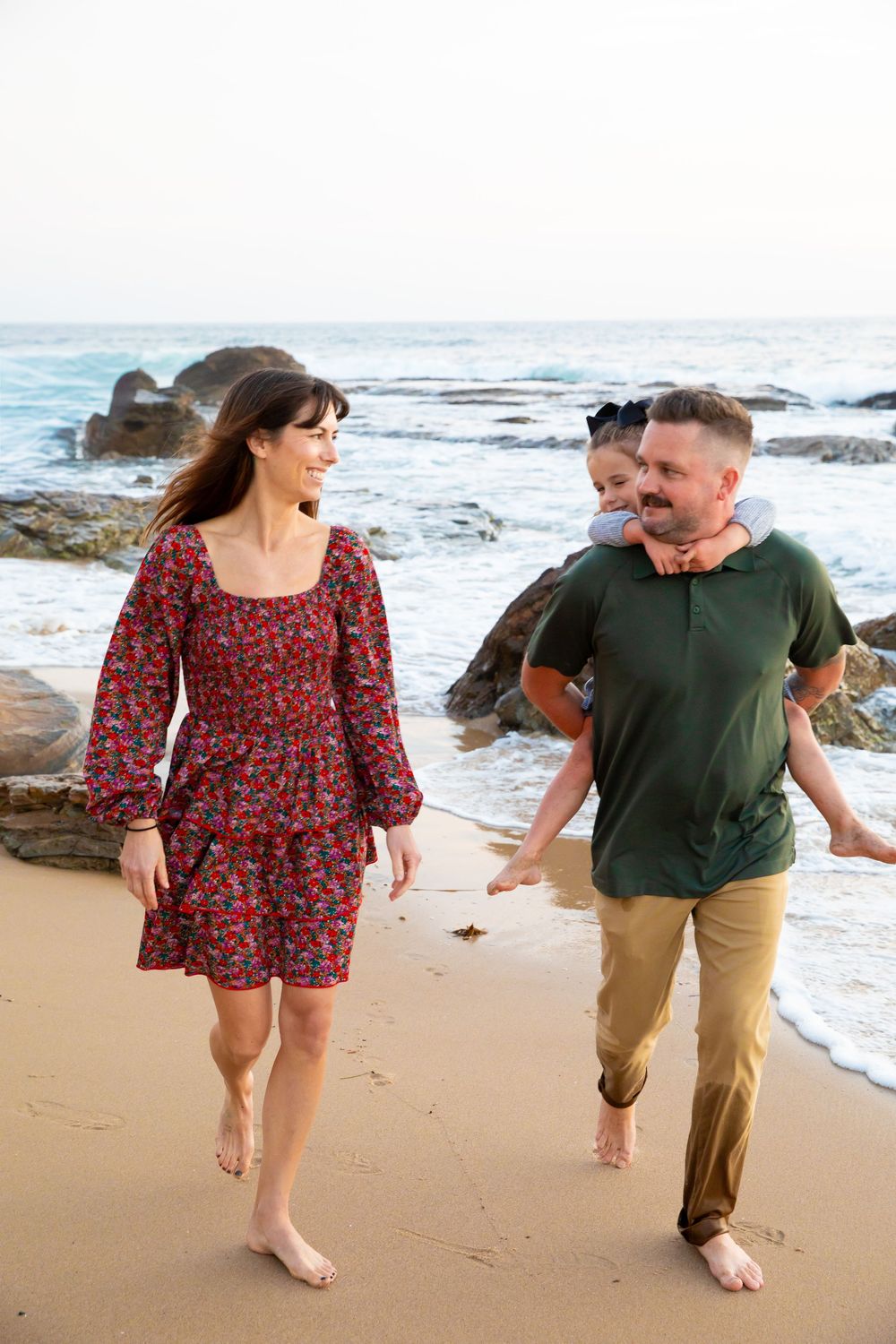 A couple walking barefoot on a beach near rocky coastline, with waves crashing in the background.
