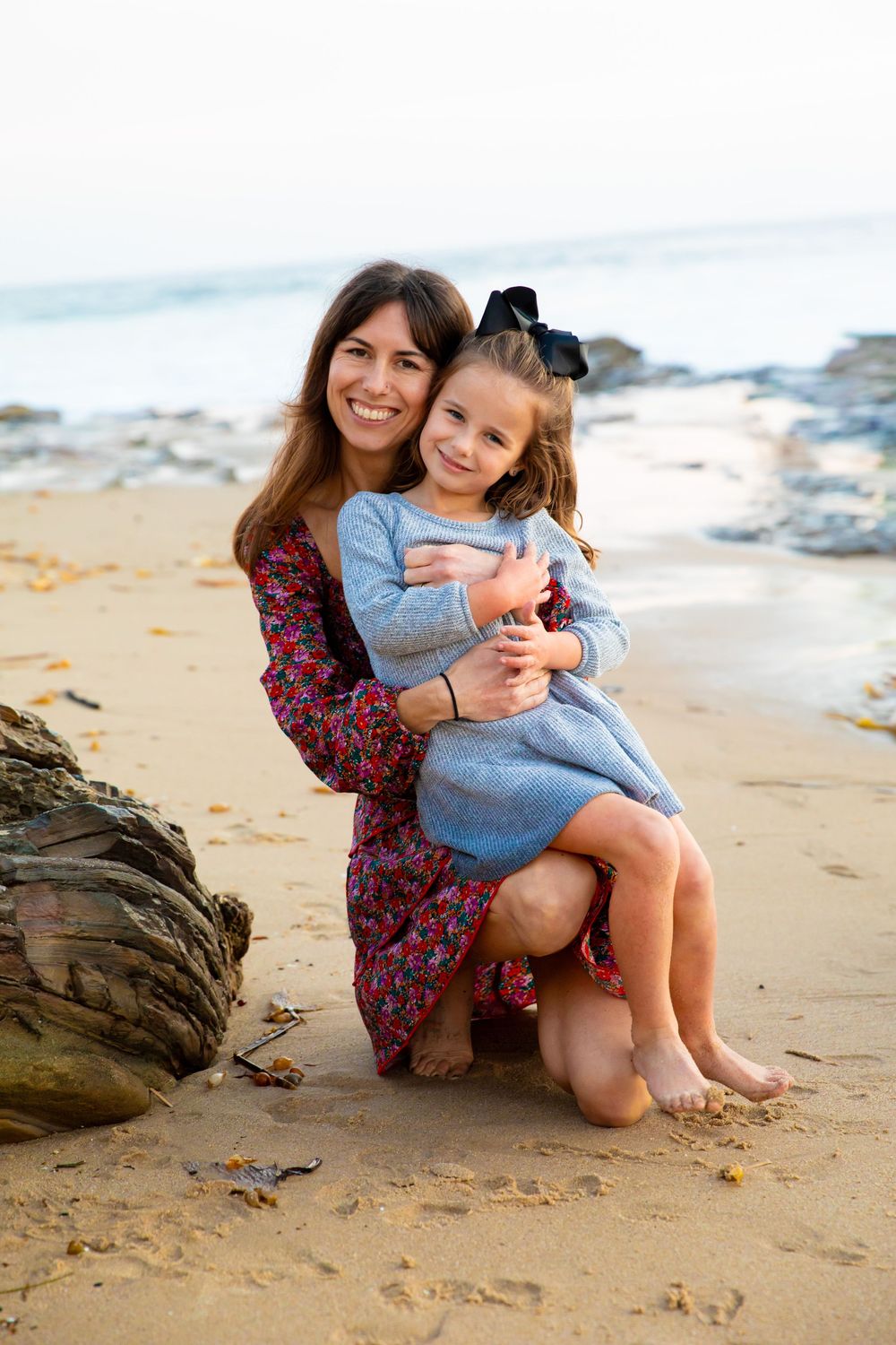 A mother and child share a tender moment while sitting together on a sandy beach near driftwood.