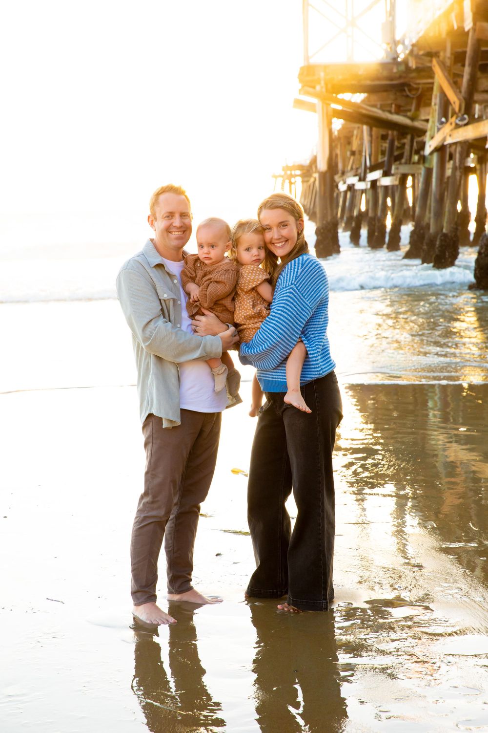 A family poses together on a sunny beach near a wooden pier with golden sunlight creating a warm backlit glow.