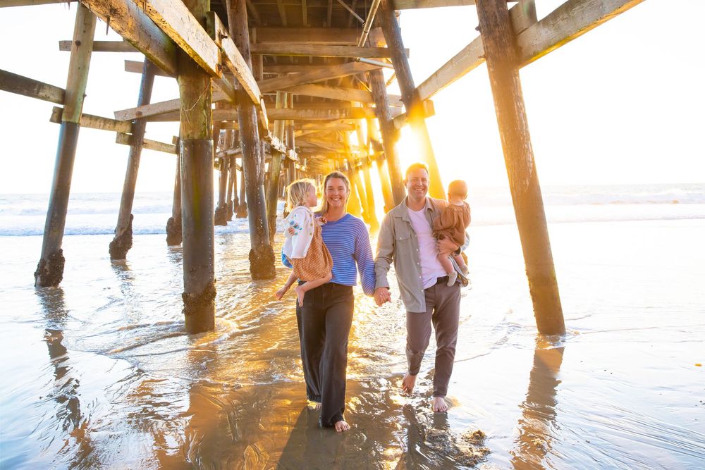 People fishing from a wooden pier at sunset with golden sunlight streaming through the pilings and reflecting on calm water.