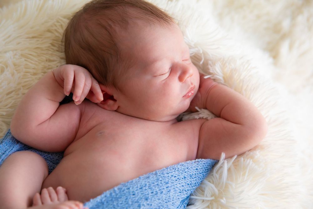 A sleeping newborn baby swaddled in a soft blue blanket rests peacefully on a white fur blanket.