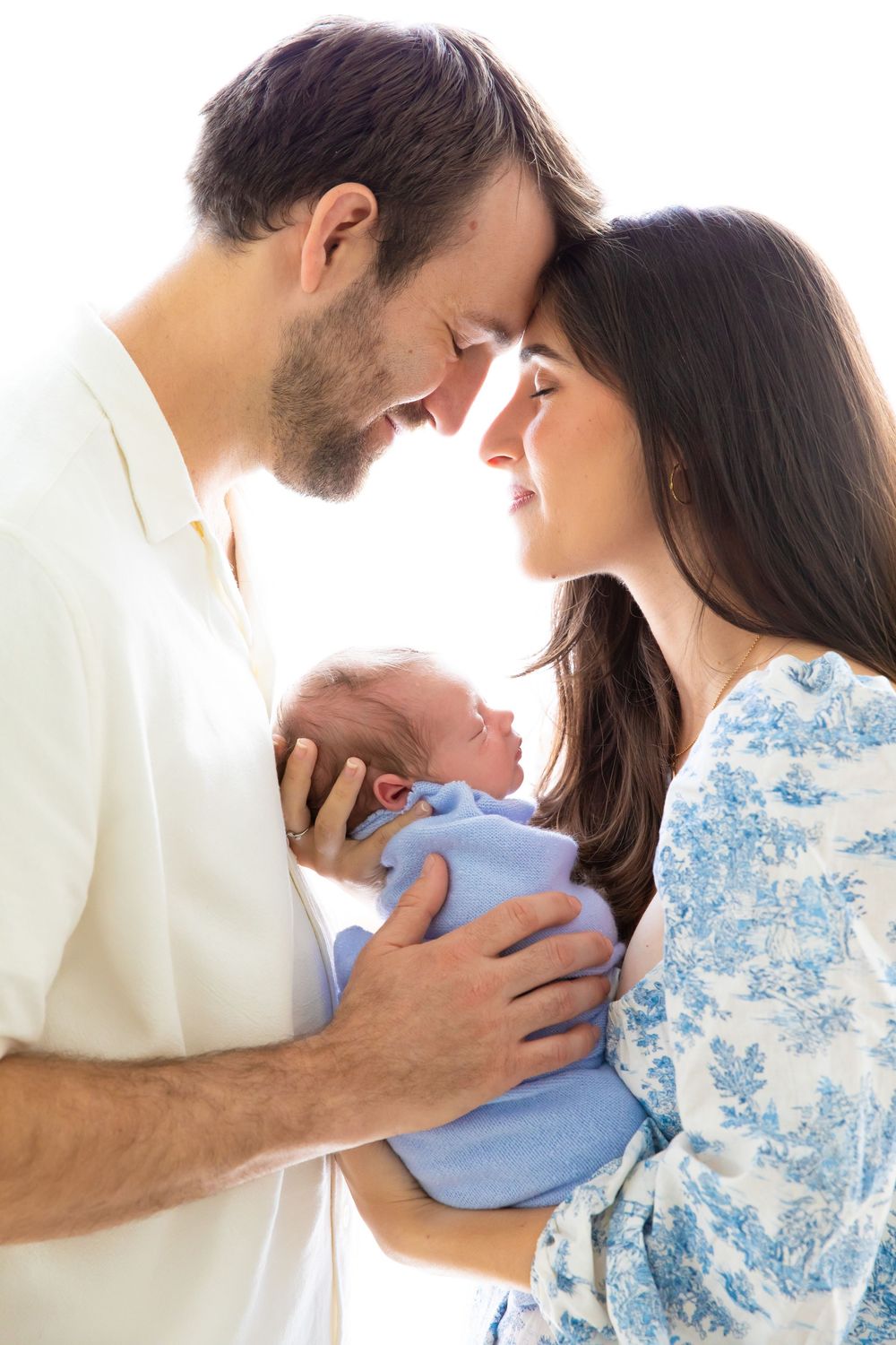 A couple in white and blue floral attire share tender moments with their newborn baby in a photo series.