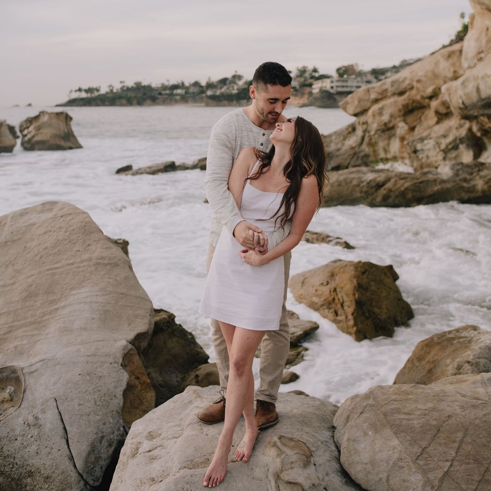 Couple embracing romantically on coastal rocks with ocean waves crashing behind them at sunset.