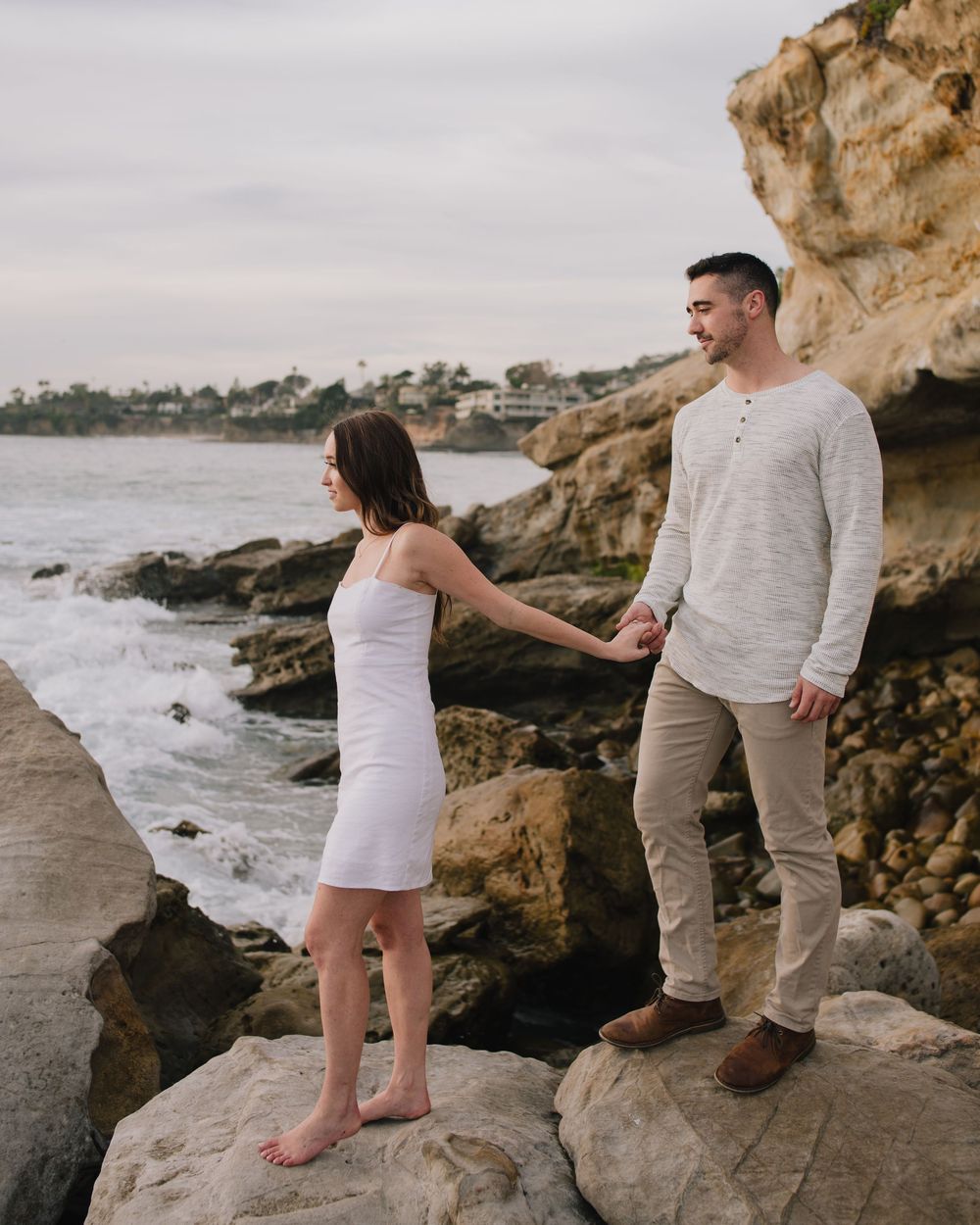 Couple holding hands on rocky coastal cliffs, woman in white dress, man in linen shirt, ocean waves in background.