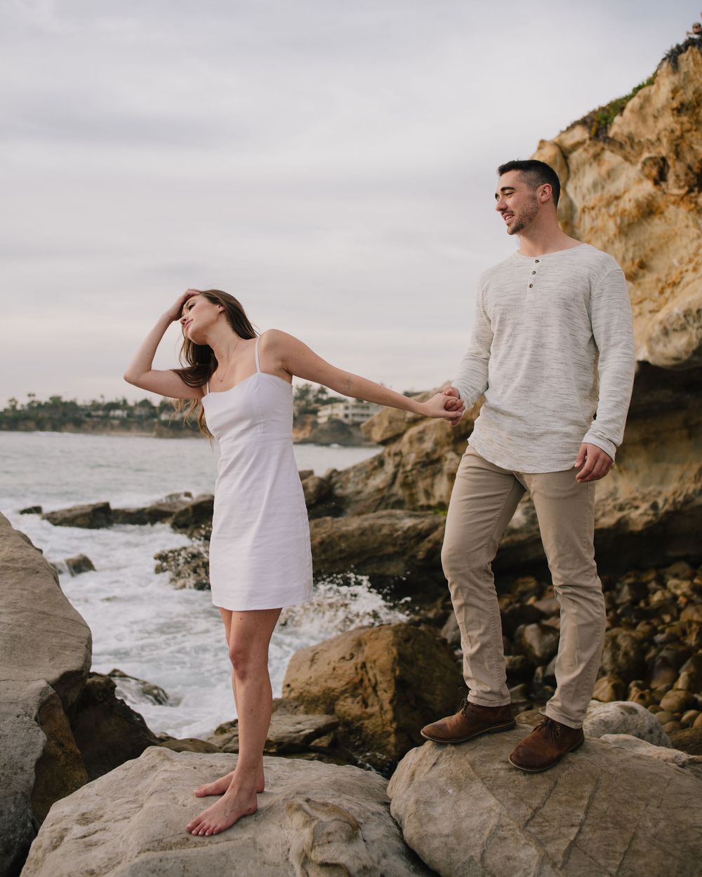 Couple holding hands on rocky coastal cliffs, woman in white dress, man in gray sweater, ocean waves behind them.