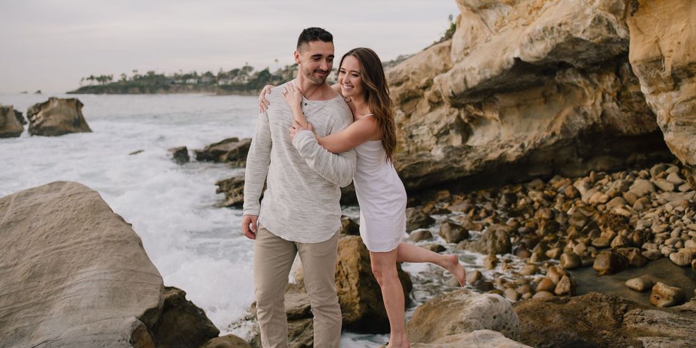 Couple embracing romantically on a rocky beach with ocean waves, man in tan suit and woman in white dress.