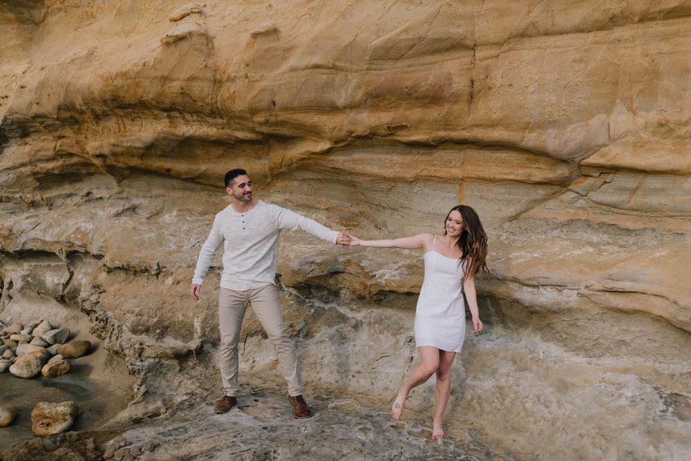 A couple holds hands and walks along a rocky sandstone cliff face during an outdoor engagement photo session.