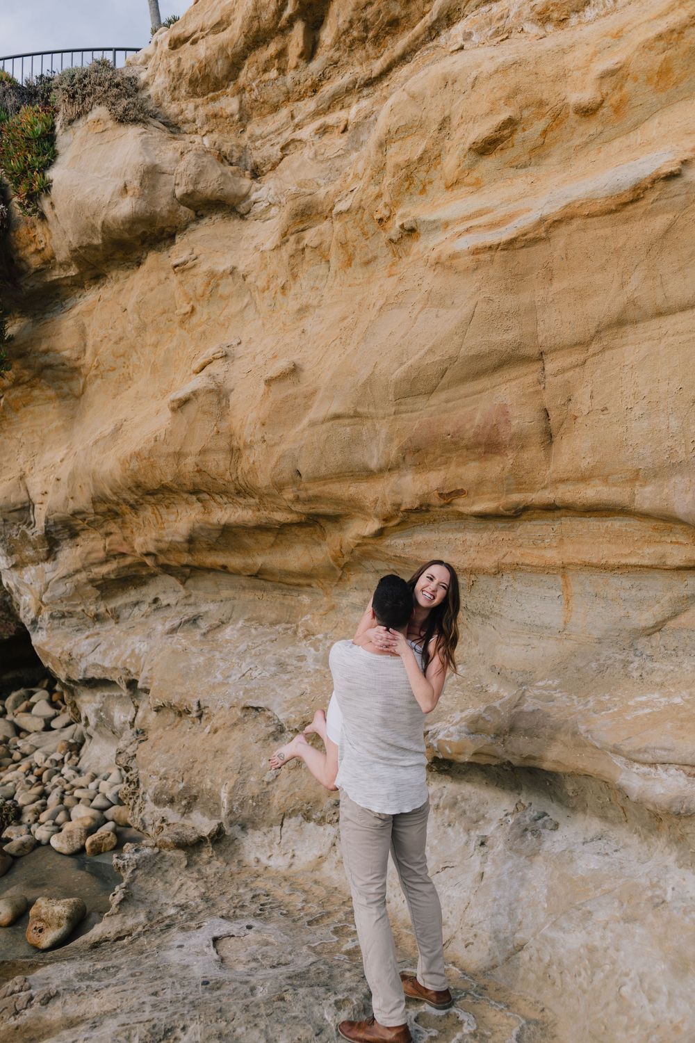 Couple embracing and laughing against golden sandstone cliffs at a rocky beach location.