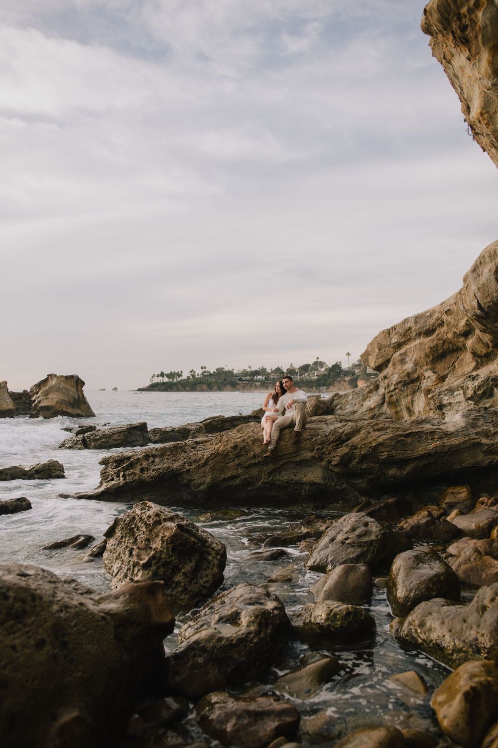 A person sits on rocky coastal cliffs overlooking the ocean waves with trees and cloudy sky in the background.