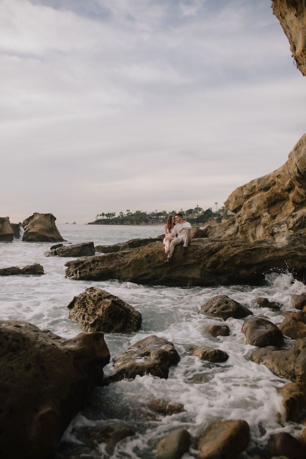Couple sitting together on rocky coastal cliffs with ocean waves crashing below at sunset.