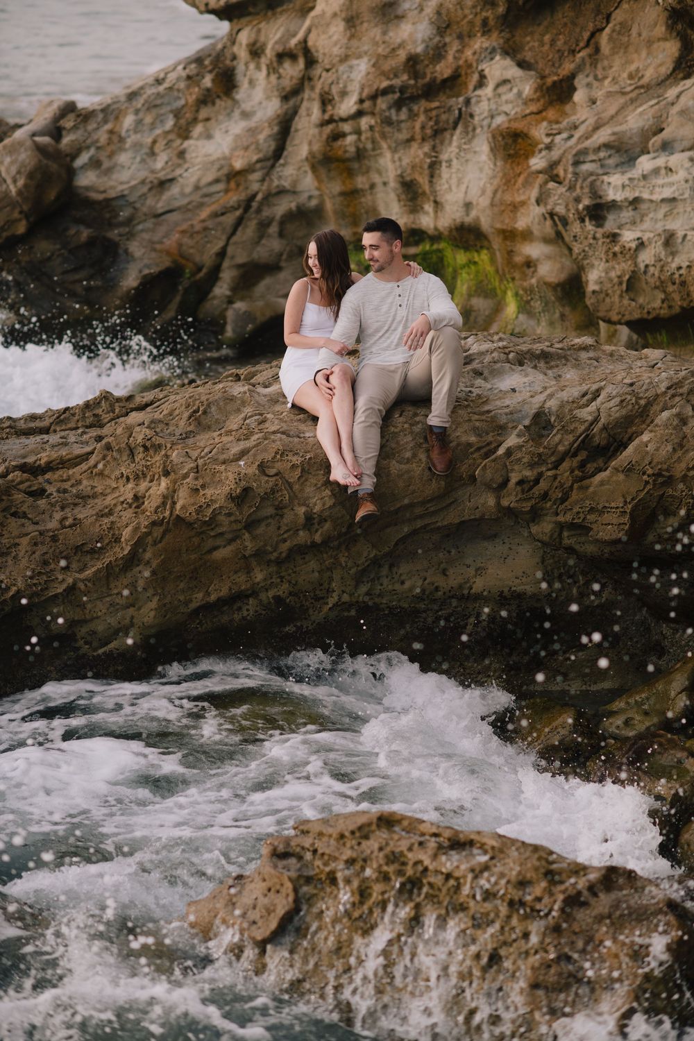 Couple sitting together on coastal rocks with ocean waves splashing below, sharing an intimate moment.