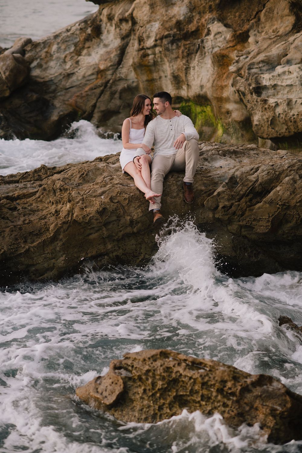 Couple sitting on rocky coastal cliffs with ocean waves crashing below, sharing a romantic moment by the sea.