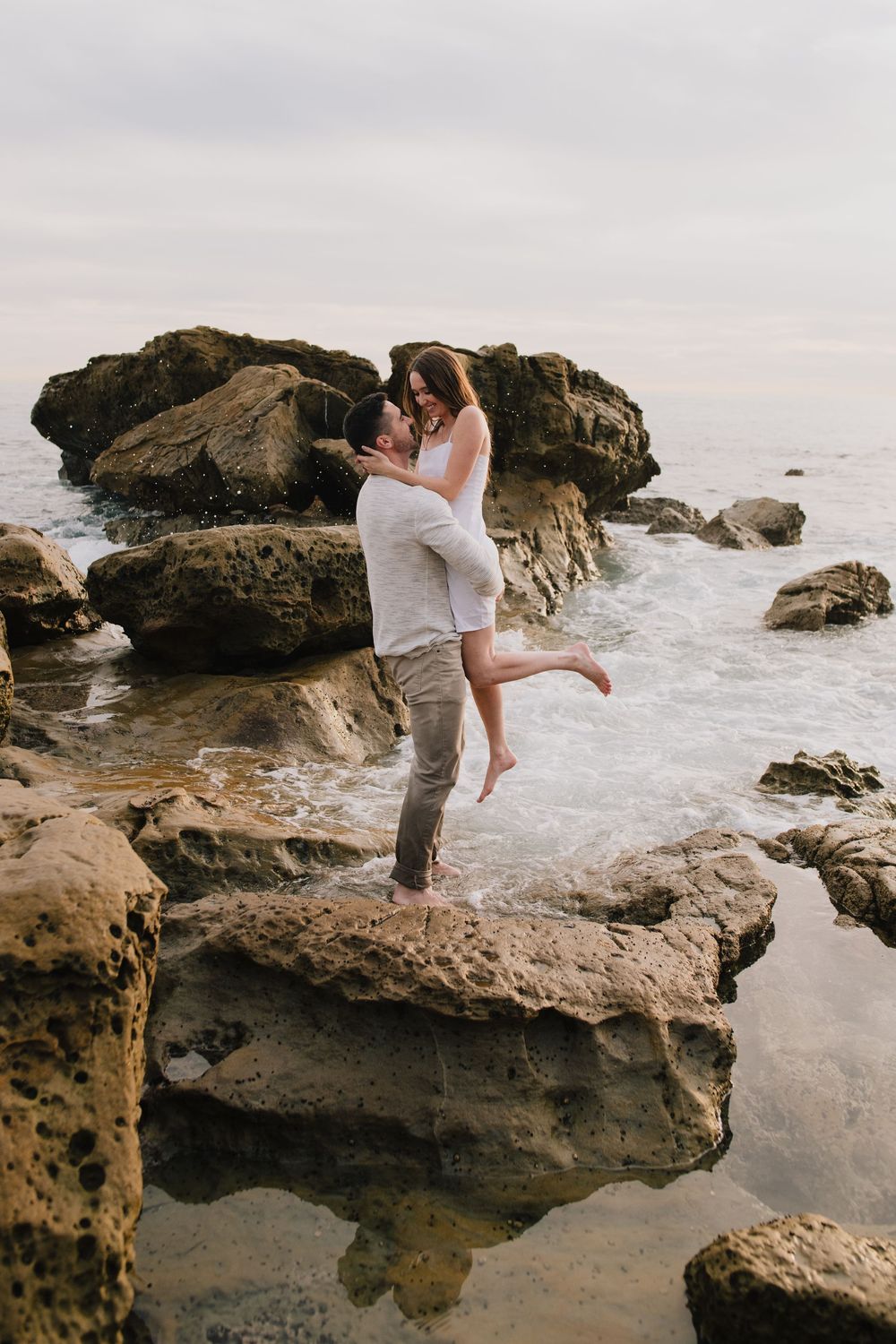 Man lifting woman on rocky seaside cliffs with ocean waves, couple sharing romantic embrace outdoors.