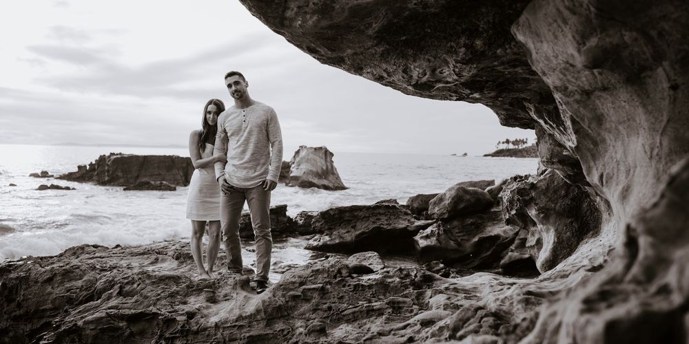 Couple holding hands under rocky arch on black and white beach with ocean waves in background.
