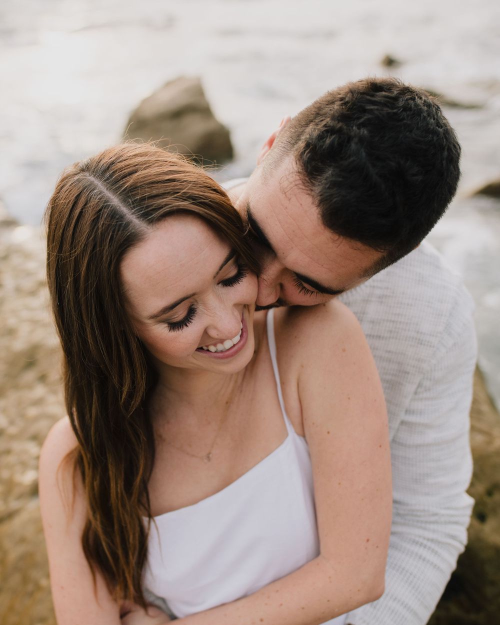 Couple sharing a tender moment on rocky beach, man nuzzling smiling woman in white dress near ocean.