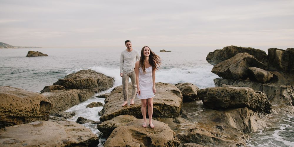 Couple standing on rocky coastal shore with ocean waves and misty sky behind them.