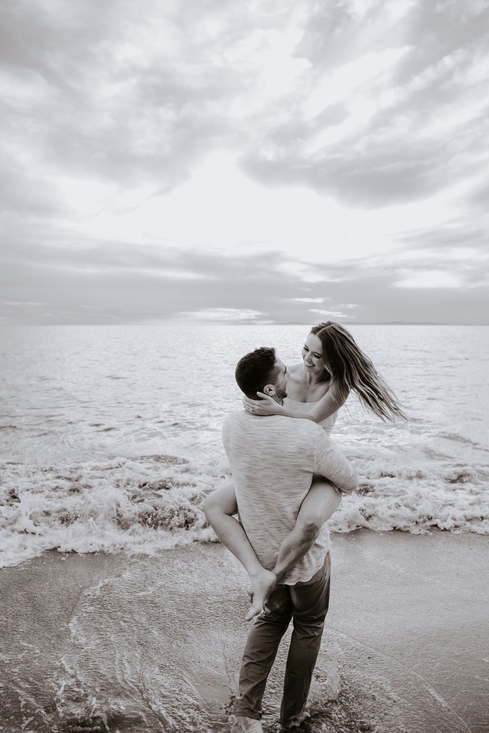 Black and white photo of a couple embracing and kissing on a beach with ocean waves and cloudy sky.