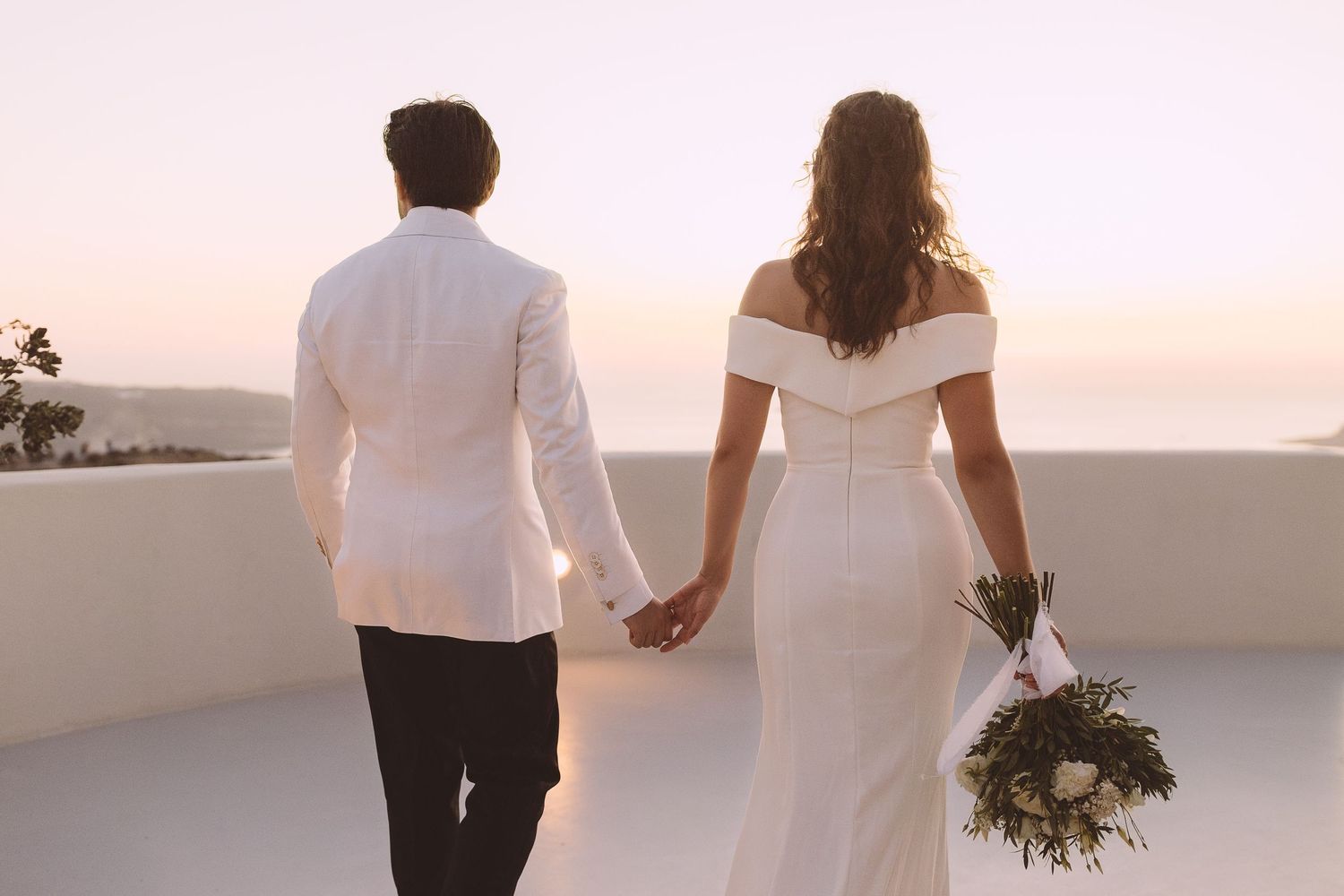 A destination wedding couple walks hand in hand at sunset overlooking a coastal view in Santorini, Greece.