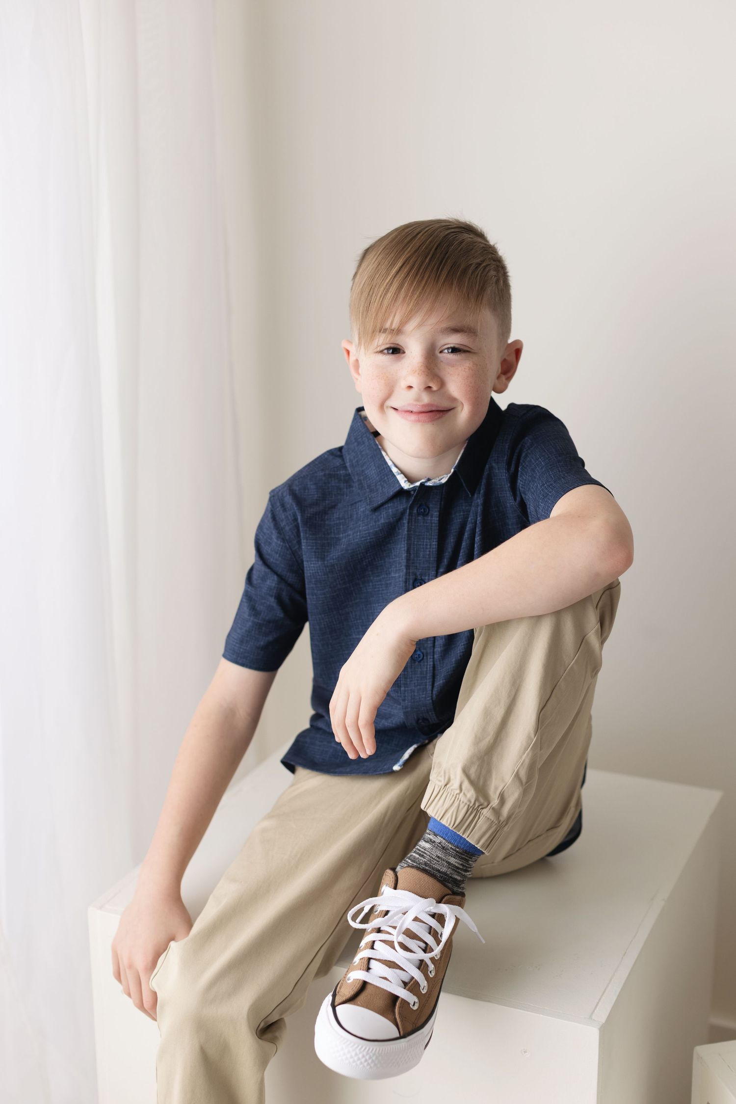 A young child in a navy shirt and khaki pants sits casually on a white surface, showing off brown and white sneakers. Elizabeth Lang Photogr