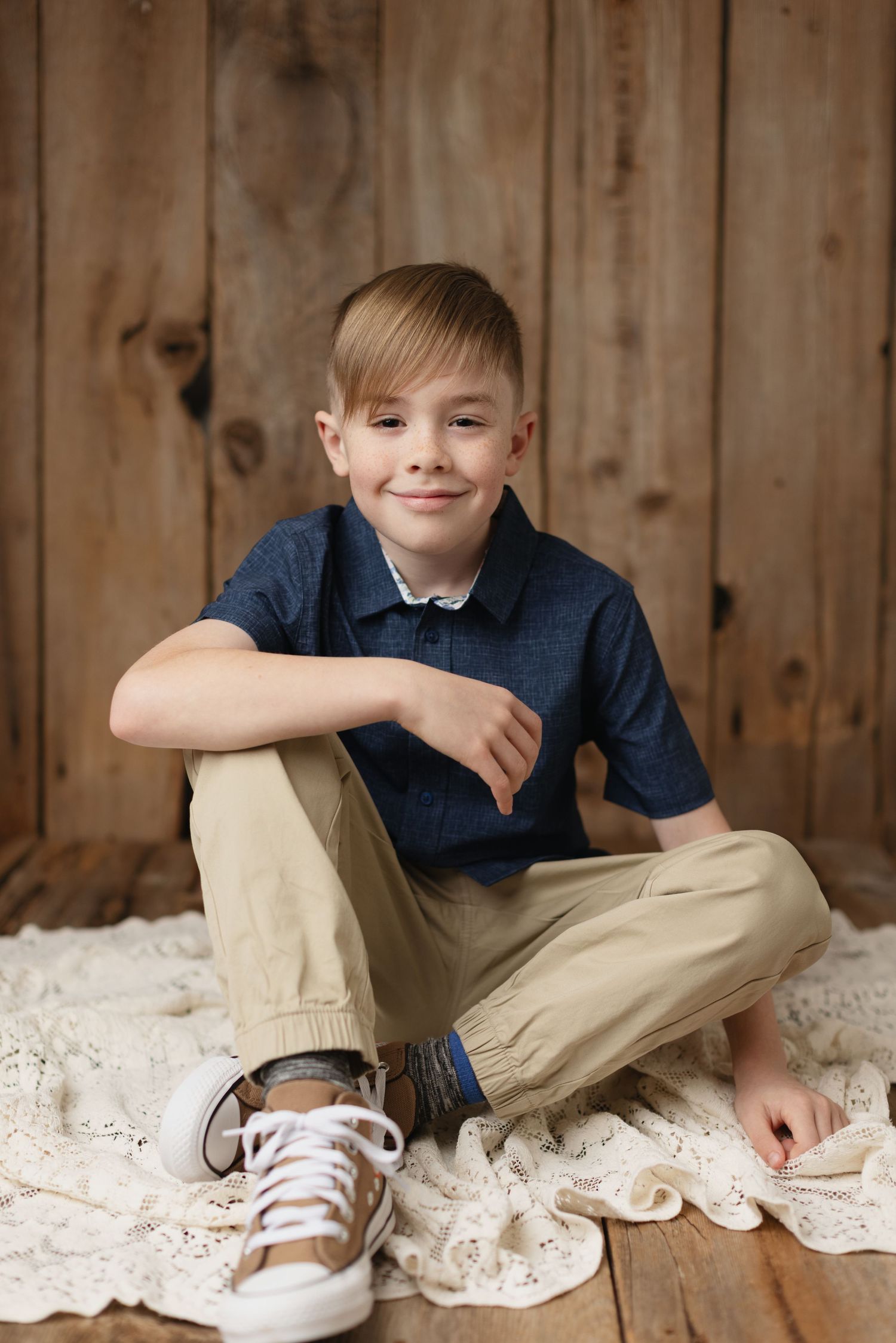 A child sits casually on a wooden floor with lace fabric, wearing khaki pants and navy shirt in a rustic setting. Elizabeth Lang Photography