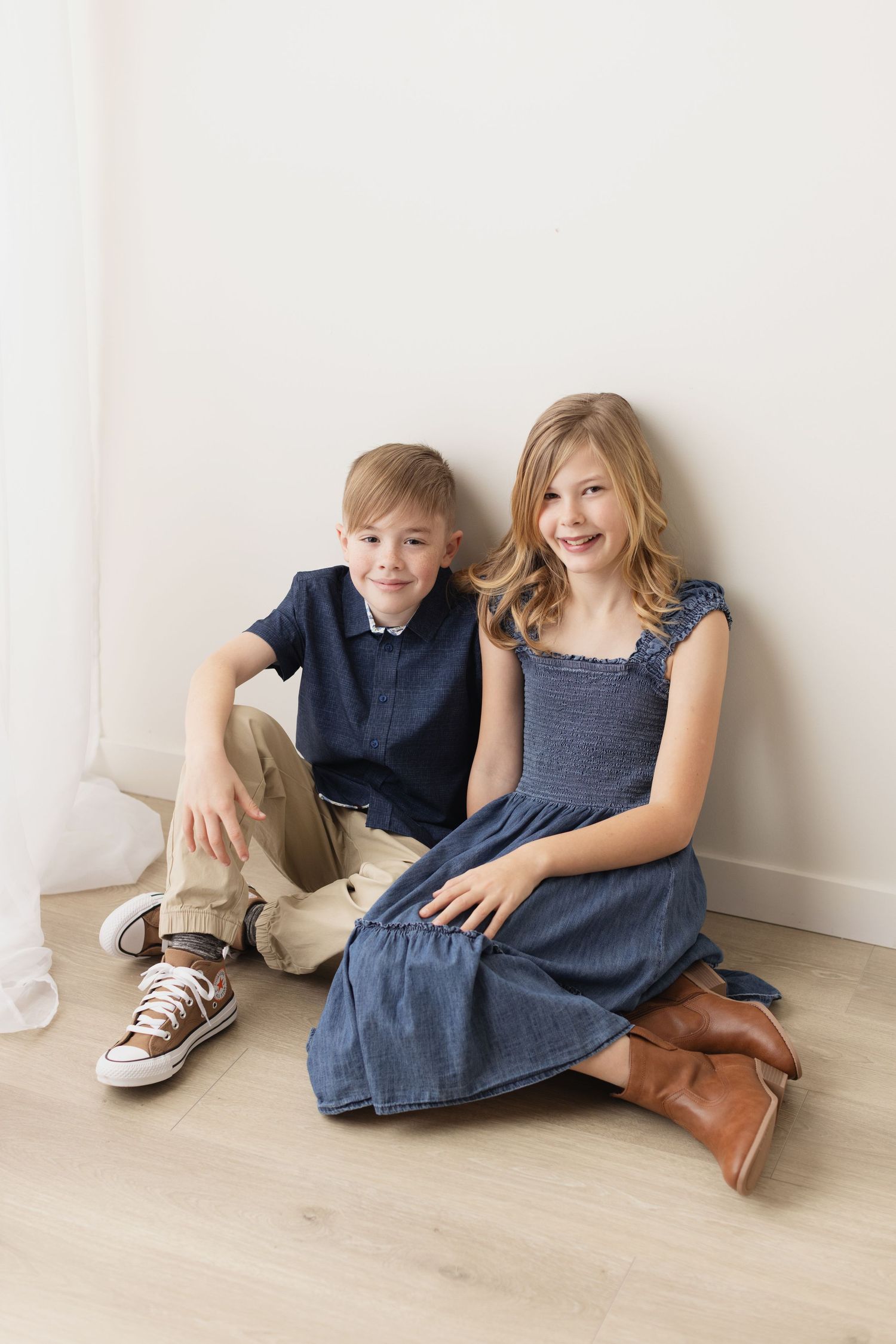 Two siblings in matching navy outfits sit casually on a light wooden floor against a white wall. Elizabeth Lang Photography Surrey