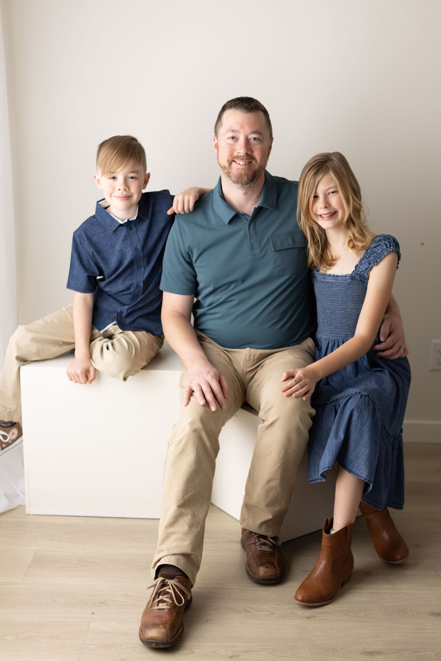 A family sits together on a white floor wearing coordinated blue and khaki outfits in a minimalist studio setting. Elizabeth Lang Photograph