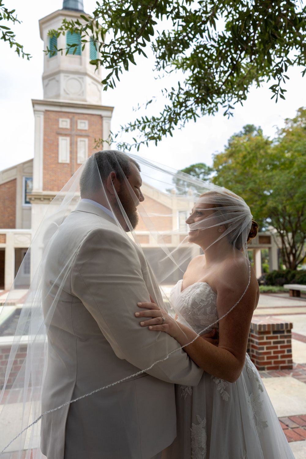 Bride and groom share romantic moment under veil near brick church bell tower.