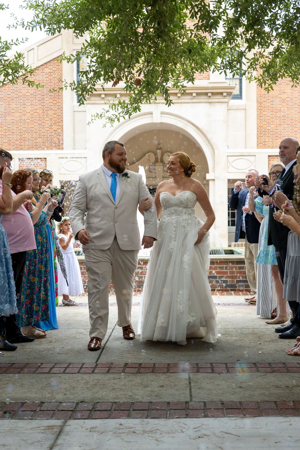 Bride and groom walk through cheering guests after outdoor church wedding ceremony.