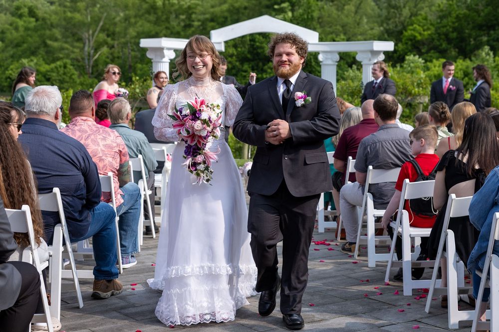 Bride in lace gown walks down aisle with groom during outdoor wedding ceremony.
