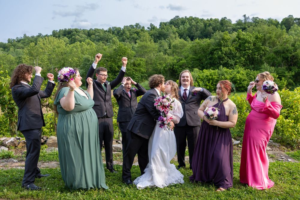 Joyful wedding party celebrating with kiss and cheers in lush green outdoor setting.