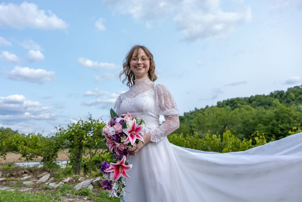 Smiling bride in white gown holding pink floral bouquet in scenic outdoor vineyard.