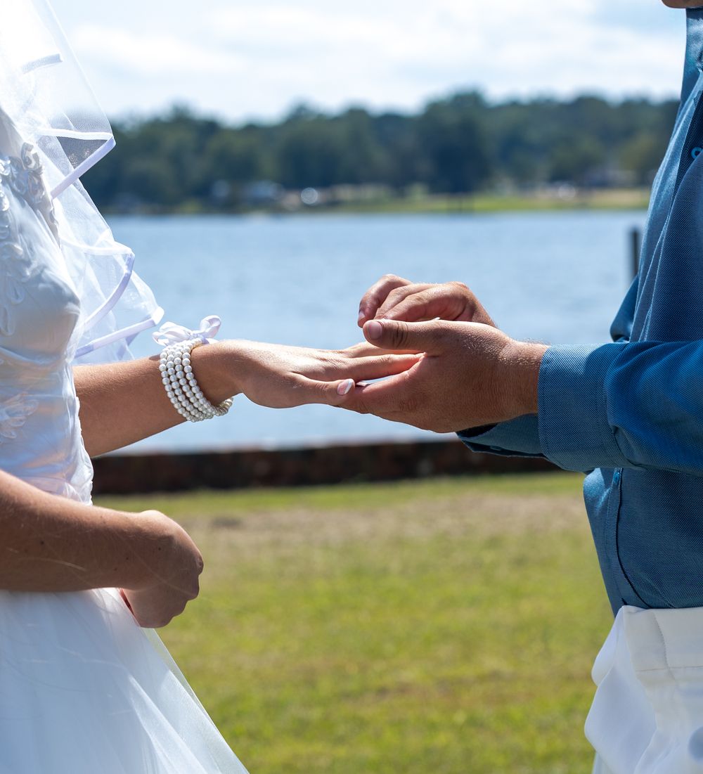 Groom places wedding ring on brides finger during outdoor lakeside ceremony.