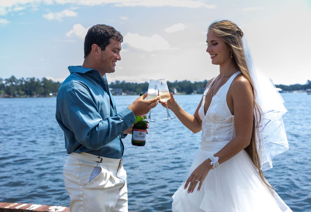 Bride and groom toasting with champagne glasses by a lakeside on their wedding day.