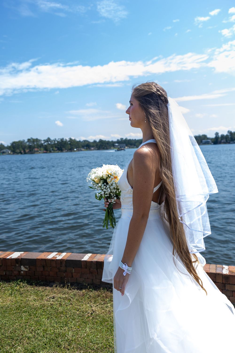 Beautiful bride in white gown holding bouquet standing by a serene lake on sunny day.