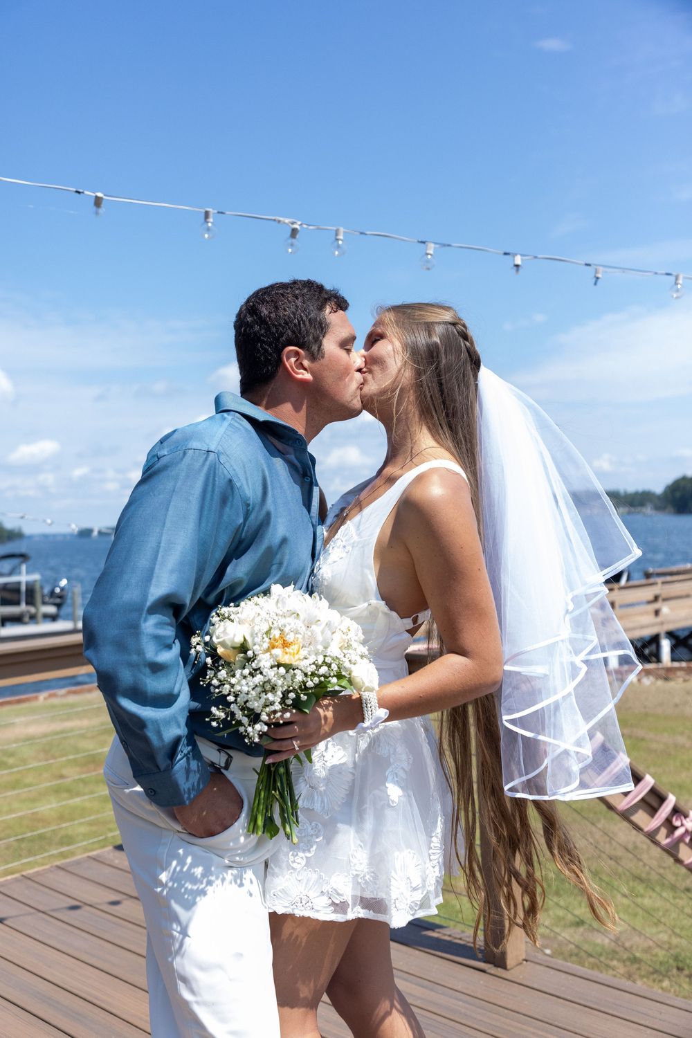 Newlywed couple sharing romantic kiss on waterfront deck with string lights overhead.