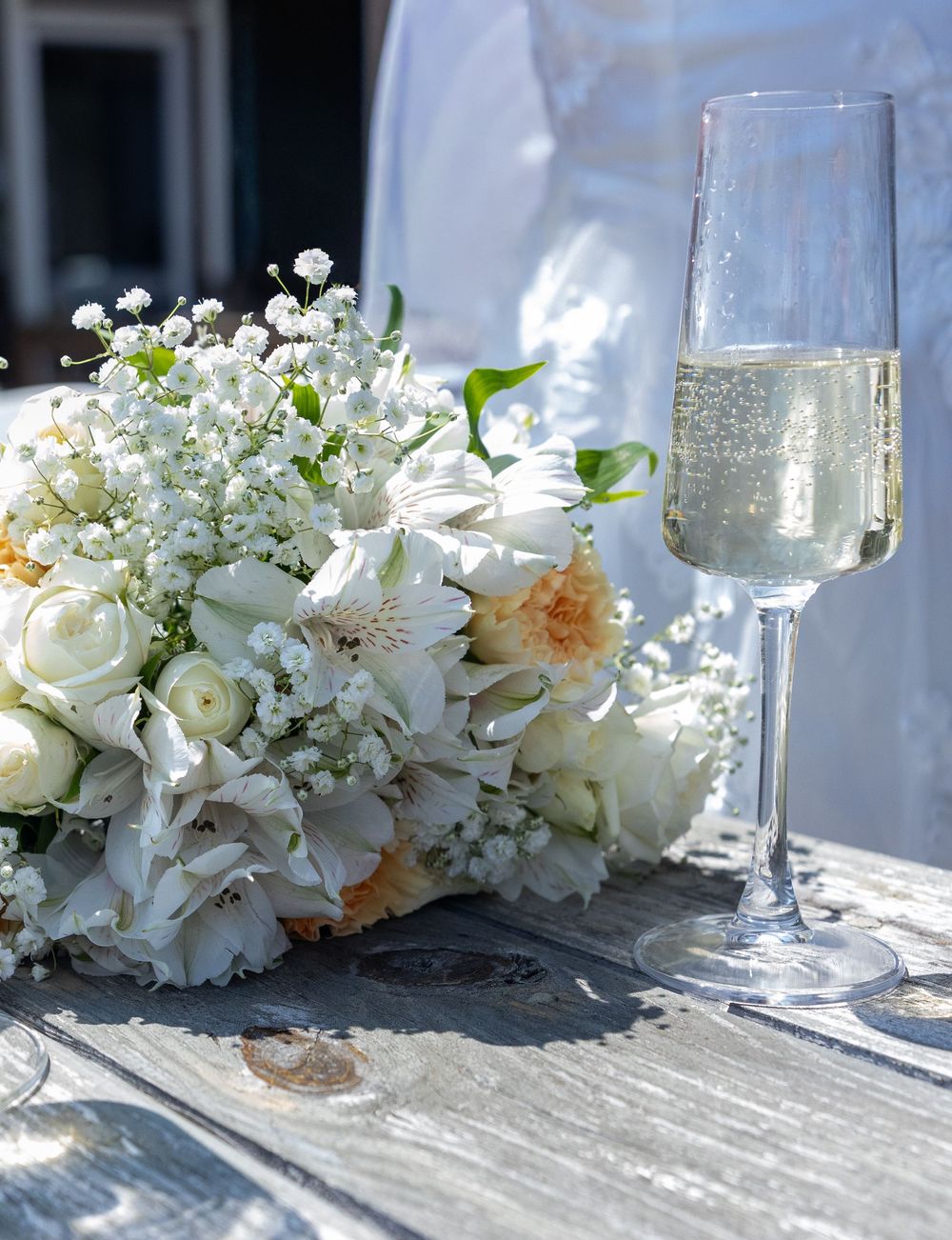 Elegant white wedding bouquet beside champagne flute on rustic wooden table outdoors.