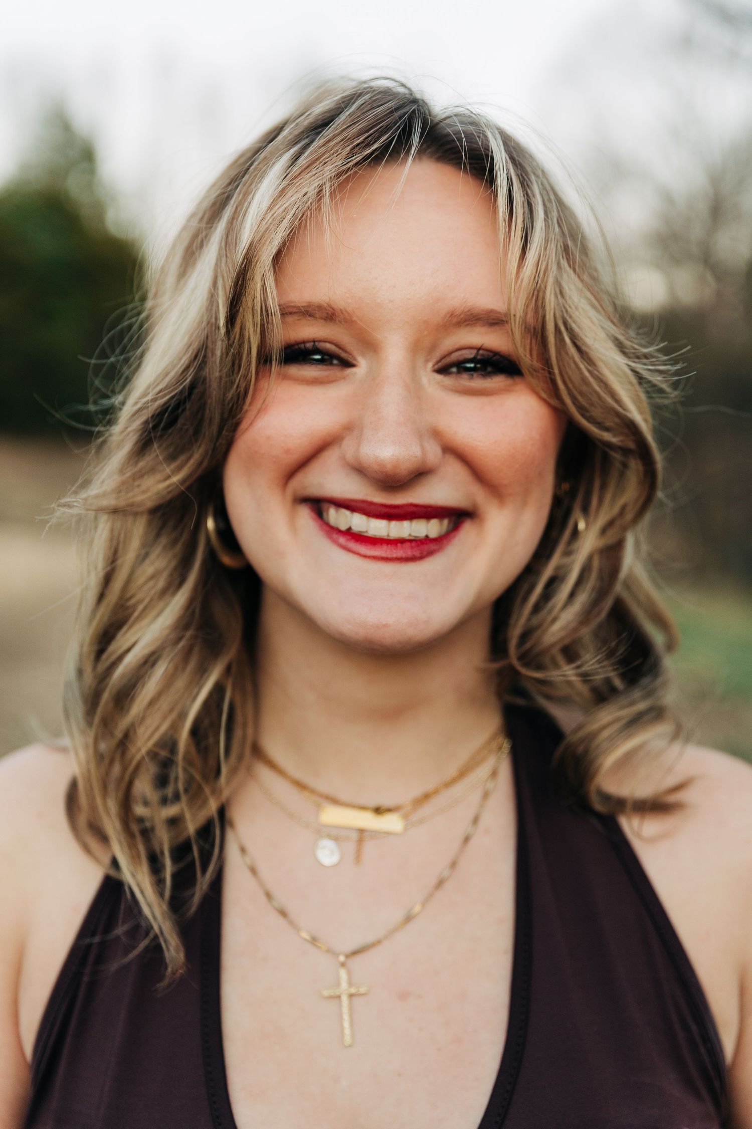Close-up portrait of someone with wavy blonde hair wearing a brown top and layered gold necklaces, smiling outdoors.