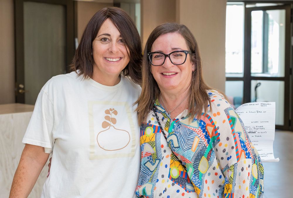 Two people standing together and smiling, one wearing a white t-shirt and one in a colorful patterned top.