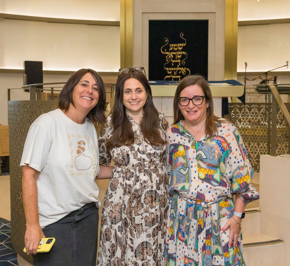 Three people pose together at an indoor event, with decorative gold elements visible in the background.