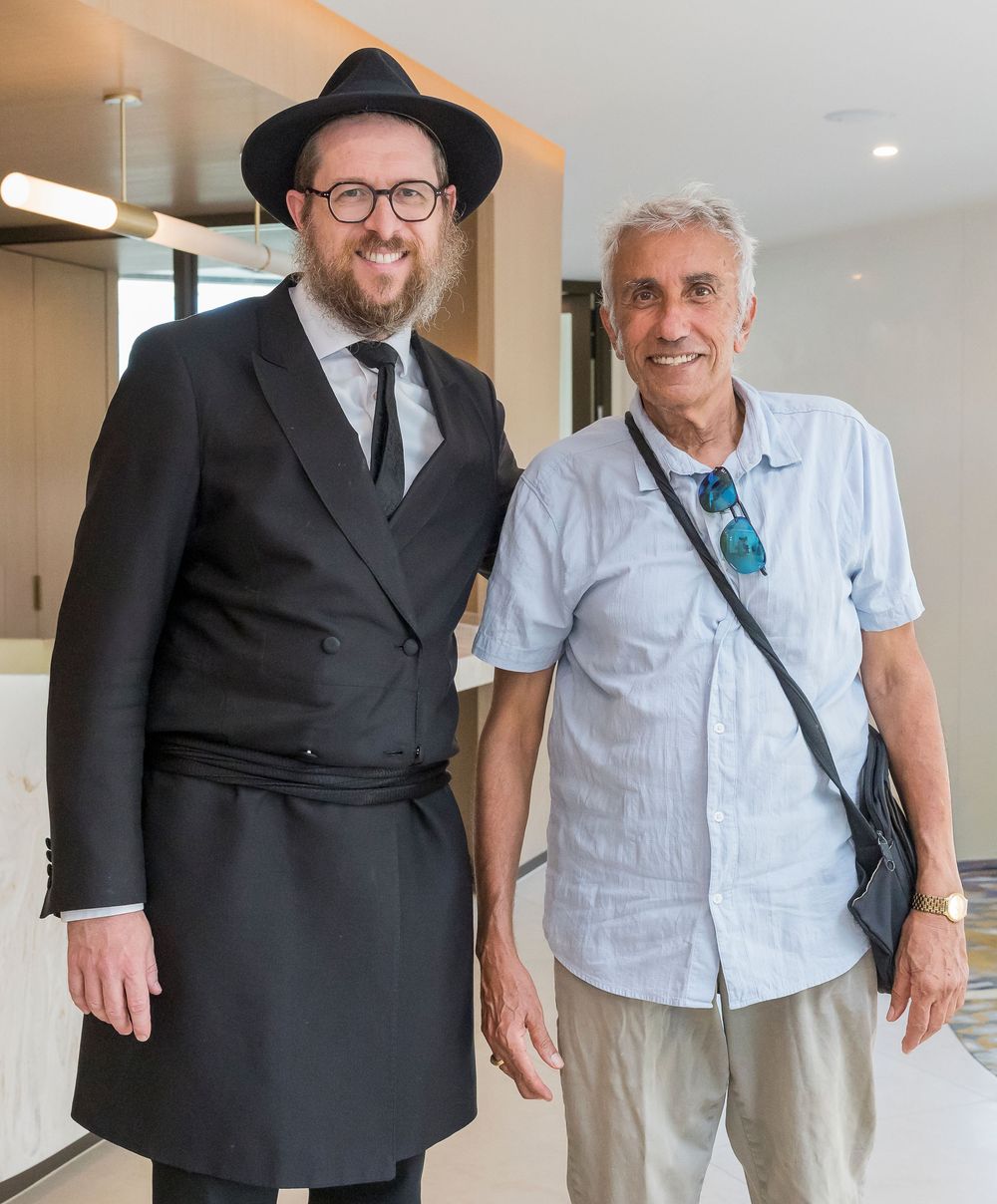 Two people standing together in a hallway, one wearing a dark suit and hat while the other wears a light shirt.